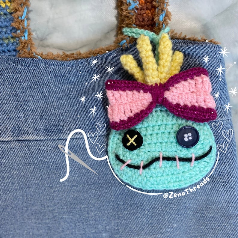 Pu&ograve; includere: Un personaggio Stitch in crochet blu turchese con un fiocco rosa e viola, capelli gialli e occhi neri. Il personaggio &egrave; attaccato a una borsa di jeans con un ago e filo bianchi.