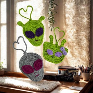 May include: Three crocheted alien-themed ornaments. Two are green with purple eyes, and one is gray with pink eyes. Each has a heart-shaped loop for hanging. The ornaments are handmade and suitable for home decor.
