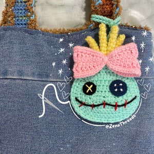 Pu&ograve; includere: Una borsa di jeans blu con un personaggio Stitch all'uncinetto attaccato alla parte anteriore. Il personaggio ha un fiocco rosa, capelli gialli e un occhio blu con una X nera. La borsa ha una tracolla in crochet marrone e stelle e cuori bianchi disegnati sulla parte anteriore. Il testo '@ZenoThreads' &egrave; scritto nell'angolo in basso a destra della borsa.