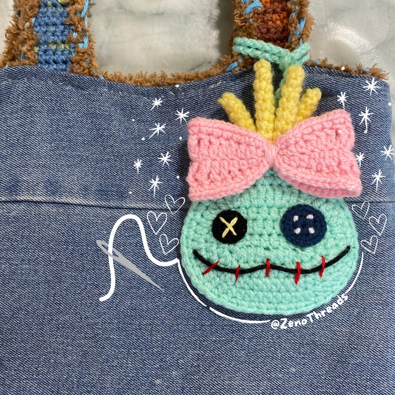 Pu&ograve; includere: Una borsa di jeans blu con un personaggio Stitch all'uncinetto attaccato alla parte anteriore. Il personaggio ha un fiocco rosa, capelli gialli e un occhio blu con una X nera. La borsa ha una tracolla in crochet marrone e stelle e cuori bianchi disegnati sulla parte anteriore. Il testo '@ZenoThreads' &egrave; scritto nell'angolo in basso a destra della borsa.