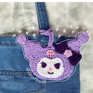 Pode incluir: Encanto de personagem de crochê roxo e branco com um detalhe de caveira e flor. O encanto está preso a uma bolsa de lona jeans. O texto "@zenoThreads" é visível na bolsa.