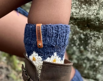 May include: Navy blue and cream-coloured knit leg warmers with a daisy pattern and a leather tag. The leg warmers are worn over a brown leather boot. The tag reads "FAIRWORKSHOP".