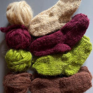 Könnte beinhalten: Eine Auswahl handgestrickter Socken in Beige-, Burgunder-, Grün- und Brauntönen, zusammen mit passenden Wollknäueln. Die Socken sind aus einem strukturierten, flauschigen Material gefertigt, und die Wollknäuel sind ordentlich gewickelt.