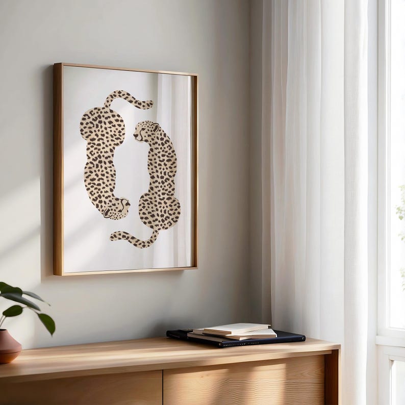 May include: A framed print of two cheetahs in a beige and brown spotted pattern, facing each other in a circular formation. The print is framed in a gold frame and is hanging on a light beige wall.