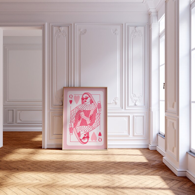 May include: A framed print of a pink Queen of Hearts playing card with a woman wearing sunglasses and holding a glass of champagne. The background is a white wall with ornate molding.