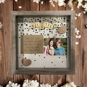 Puede incluir: Una caja de sombra enmarcada con fondo blanco y corazones negros. La palabra "Hermana" está deletreada con fichas de letras de madera. Una foto de dos mujeres está en la caja, y una nota escrita a mano dice "Lado a lado o a kilómetros de distancia, somos hermanas, conectadas por el corazón."