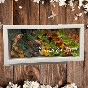 May include: A white frame with a glass front containing pressed flowers in shades of orange, yellow, and green. The text "Cecelia & Mark 10-30-22" is printed in white on the glass.