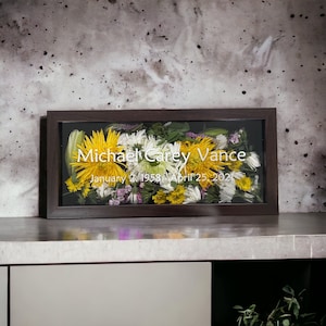 May include: A dark brown wooden frame with a glass front displays pressed flowers and foliage. The text "Michael Carey Vance" and "January 6, 1958 - April 25, 2021" is printed in white on the glass.
