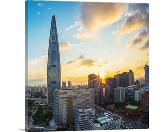 May include: A tall, modern skyscraper with a glass facade stands out against a backdrop of a city skyline at sunset. The sky is a vibrant mix of blue, orange, and pink, with fluffy clouds. The sun is setting behind the building, casting a warm glow on the city.