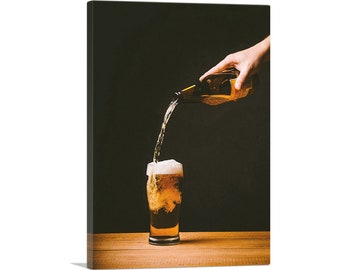 May include: A hand pours beer from a brown bottle into a clear glass with a white head of foam. The glass is on a wooden table.