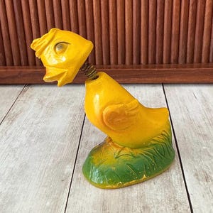 May include: A vintage yellow bird-shaped bottle opener with a spring neck. The bird is perched on a green and yellow base with painted grass details. The opener is made of a painted material and has a detailed head.