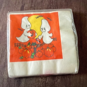 May include: A white paper napkin with a Halloween theme. The napkin features two ghosts holding a jack-o-lantern, a black cat, and orange pumpkins on a red background.