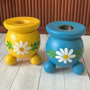 May include: Two hand-painted wooden candle holders. One is yellow, the other is blue. Each has a white daisy design with green leaves. They have a round base with three small ball feet. The candle holders are decorative.