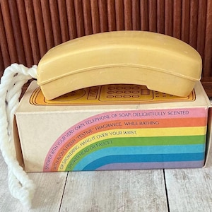 May include: A vintage yellow telephone-shaped soap bar with a white rope attached, resting on a box with a rainbow design and text. The box reads "Shower with your very own telephone of soap."