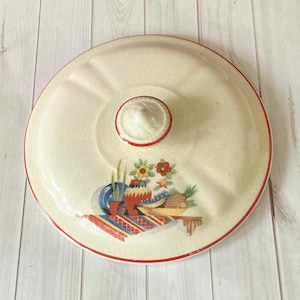 May include: Cream ceramic lid with a red trim and a central knob. The lid features a colourful illustration of a still life with flowers, fruit, and pottery. The design includes a red, blue, yellow, and green colour palette.