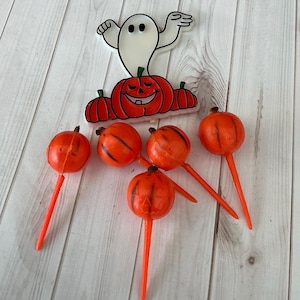 May include: A collection of Halloween decorations featuring a smiling jack-o'-lantern, a friendly ghost, and several orange pumpkin-shaped picks. The decorations are set against a light wood grain background.
