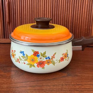 May include: A vintage enamelware saucepan with a lid. The lid is orange and yellow with a brown handle. The white pot is decorated with a floral pattern in red, yellow, blue, and orange. The handle is brown.
