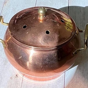 May include: A copper pot with a lid and brass handles. The pot has a round shape with a lid that has three small holes. The lid is topped with a brass knob. The pot is sitting on a wooden surface.