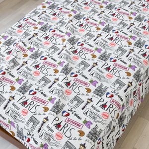 May include: White fabric with a repeating pattern of Parisian landmarks, including the Eiffel Tower and Arc de Triomphe, along with the word "PARIS" and phrases like "Bonjour" and "Je t'aime". The design also features stamps, hearts, and grapes.