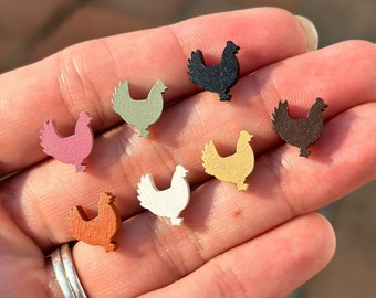 May include: A set of eight wooden chicken silhouette earrings in various colours, including black, white, brown, pink, green, and yellow. The earrings are displayed on a hand.