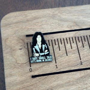 Pode incluir: Um alfinete de esmalte preto e branco com uma figura estilizada de cabelo longo e escuro e uma jaqueta. O alfinete tem o texto "I HATE SMALL TALK LET'S DRINK IN SILENCE". O alfinete est&aacute; em uma superf&iacute;cie de madeira com uma r&eacute;gua.