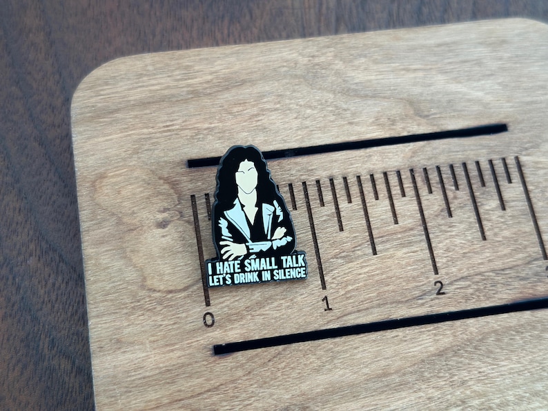 Pode incluir: Um alfinete de esmalte preto e branco com uma figura estilizada de cabelo longo e escuro e uma jaqueta. O alfinete tem o texto "I HATE SMALL TALK LET'S DRINK IN SILENCE". O alfinete est&aacute; em uma superf&iacute;cie de madeira com uma r&eacute;gua.