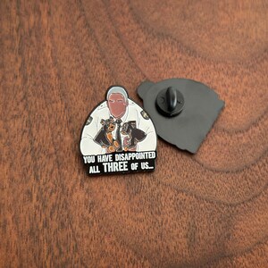 Pode incluir: Um pin de esmalte preto e branco com uma imagem de desenho animado de um homem em uniforme de pol&iacute;cia com dois c&atilde;es. O texto "YOU HAVE DISAPPOINTED ALL THREE OF US..." est&aacute; abaixo. O pin est&aacute; em uma superf&iacute;cie de madeira.