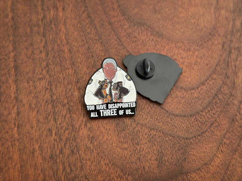 Pode incluir: Um pin de esmalte preto e branco com uma imagem de desenho animado de um homem em uniforme de pol&iacute;cia com dois c&atilde;es. O texto "YOU HAVE DISAPPOINTED ALL THREE OF US..." est&aacute; abaixo. O pin est&aacute; em uma superf&iacute;cie de madeira.