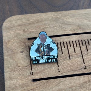 Pode incluir: Pin de esmalte com uma pessoa em uniforme de pol&iacute;cia com dois c&atilde;es. O pin tem o texto "YOU HAVE DISAPPOINTED ALL THREE OF US...". O pin est&aacute; em uma superf&iacute;cie de madeira com marca&ccedil;&otilde;es de r&eacute;gua.