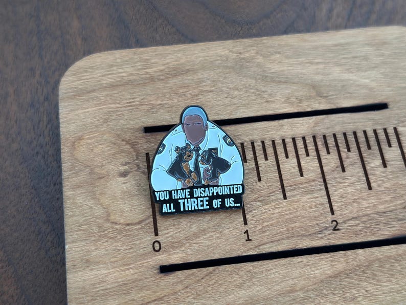Pode incluir: Pin de esmalte com uma pessoa em uniforme de pol&iacute;cia com dois c&atilde;es. O pin tem o texto "YOU HAVE DISAPPOINTED ALL THREE OF US...". O pin est&aacute; em uma superf&iacute;cie de madeira com marca&ccedil;&otilde;es de r&eacute;gua.