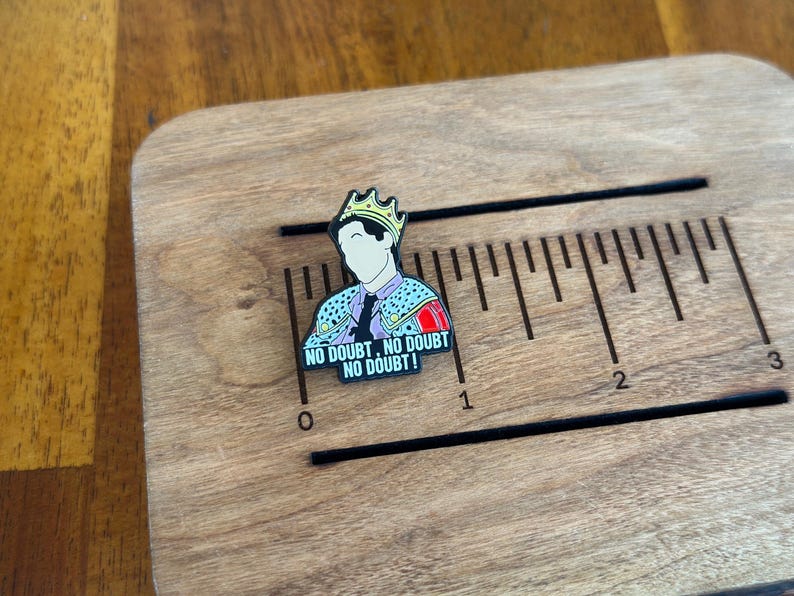 Pode incluir: Um alfinete de esmalte colorido com uma figura de desenho animado usando uma coroa e uma jaqueta estampada. O alfinete tem o texto "NO DOUBT, NO DOUBT, NO DOUBT!" e &eacute; colocado em uma r&eacute;gua de madeira. A r&eacute;gua tem aproximadamente 7,6 cm de comprimento.