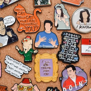 Könnte beinhalten: Eine Sammlung von Emaille-Pins mit Figuren und Zitaten aus der beliebten Fernsehserie "Friends". Die Pins sind bunt und zeigen verschiedene Designs, darunter eine Katze, einen Bilderrahmen und eine Figur, die "Ich bin gut" sagt.