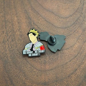 Pode incluir: Pin de esmalte com uma figura de desenho animado usando uma coroa de ouro e uma jaqueta decorativa. O pin inclui o texto "NO DOUBT, NO DOUBT, NO DOUBT!" em letras brancas sobre um fundo preto. O pin tem um fecho borboleta preto.