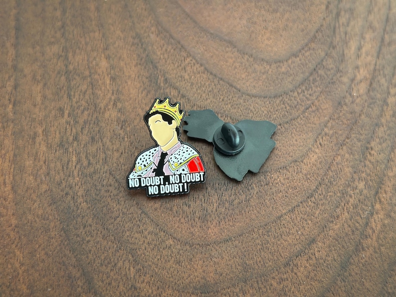 Pode incluir: Pin de esmalte com uma figura de desenho animado usando uma coroa de ouro e uma jaqueta decorativa. O pin inclui o texto "NO DOUBT, NO DOUBT, NO DOUBT!" em letras brancas sobre um fundo preto. O pin tem um fecho borboleta preto.