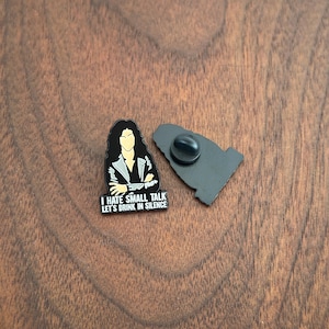 Pode incluir: Um alfinete de esmalte preto e branco com uma ilustra&ccedil;&atilde;o estilizada de uma pessoa com cabelo comprido e o texto "I HATE SMALL TALK LET'S DRINK IN SILENCE". O alfinete est&aacute; ao lado do seu suporte preto.