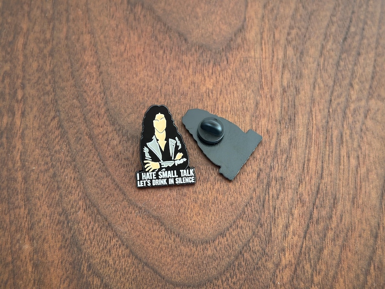 Pode incluir: Um alfinete de esmalte preto e branco com uma ilustra&ccedil;&atilde;o estilizada de uma pessoa com cabelo comprido e o texto "I HATE SMALL TALK LET'S DRINK IN SILENCE". O alfinete est&aacute; ao lado do seu suporte preto.