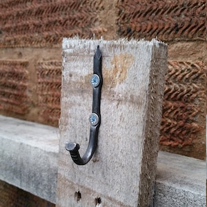 May include: A black metal hook is attached to a wooden board with two screws. The hook is shaped like a J and has a pointed top.