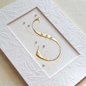May include: A white card with a gold calligraphy letter S in a white frame with a leaf pattern. The letter S is decorated with small rhinestones.