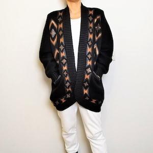 Op de afbeelding: Een zwarte gebreide cardigan met een geometrisch patroon in bruin en oranje. De cardigan heeft een shawlkraag en twee zakken.