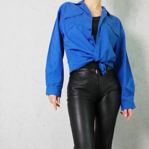 Pode incluir: Uma mulher vestindo cal&ccedil;as de couro preto e uma camisa azul brilhante abotoada amarrada na cintura.