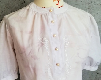 May include: A white, short-sleeved blouse with a round neckline and a row of pearl buttons down the front. The blouse features delicate white embroidery of floral designs.