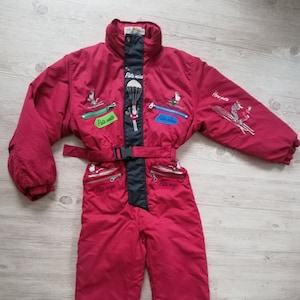 Könnte beinhalten: Roter Skianzug mit schwarzen Akzenten und Stickereien. Der Anzug hat eine Kapuze, einen Reißverschluss und einen Gürtel. Der Anzug hat mehrere Taschen und der Text "Pito nieve" ist auf der Vorderseite gestickt.