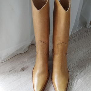 K&ouml;nnte beinhalten: Ein Paar hellbraune Leder-Cowboystiefel. Die Stiefel haben eine spitze Zehenpartie, einen hohen Schaft und einen V-f&ouml;rmigen oberen Abschluss. Die Stiefel sind aus glattem Leder gefertigt und haben ein klassisches Western-Design.