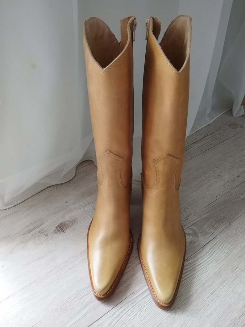 K&ouml;nnte beinhalten: Ein Paar hellbraune Leder-Cowboystiefel. Die Stiefel haben eine spitze Zehenpartie, einen hohen Schaft und einen V-f&ouml;rmigen oberen Abschluss. Die Stiefel sind aus glattem Leder gefertigt und haben ein klassisches Western-Design.