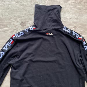 Puede incluir: Una camiseta negra de manga larga con cuello alto y el logotipo de FILA bordado en rojo, blanco y azul en la parte superior de la espalda. Las mangas presentan una cinta negra con el nombre de la marca FILA en blanco y detalles rojos.