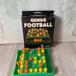 May include: A green and yellow Genius Football game board with orange and yellow pegs. The game is in its original box, which is black with white text that reads "Genius Football".