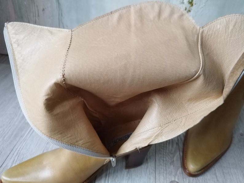 K&ouml;nnte beinhalten: Hellbraune Lederstiefel mit spitzer Zehenpartie und Blockabsatz. Die Stiefel haben einen Rei&szlig;verschluss und ein helles Innenfutter. Das Leder hat einen leichten Glanz, und die Stiefel scheinen ungetragen zu sein. Die Stiefel sind neutral, passend zu verschiedenen Outfits.