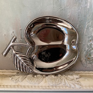 Może przedstawiać: Srebrna miska w kształcie jabłka z wzorem liścia i uchwytem. Miska jest wykonana z metalu i ma błyszczące wykończenie.