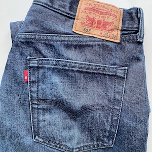 May include: Dark blue denim jeans with a classic five-pocket design. The jeans feature a red Levi's tag on the back pocket and a leather patch with the Levi Strauss & Co. logo. The waist size is W34 and the length is 81 cm.