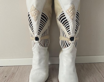 Peut inclure: Une paire de bottes blanches arrivant au genou avec un bout pointu et une tige structurée. La partie supérieure présente un détail carré tressé, un anneau central et des découpes verticales avec des bandes superposées blanches et noires.